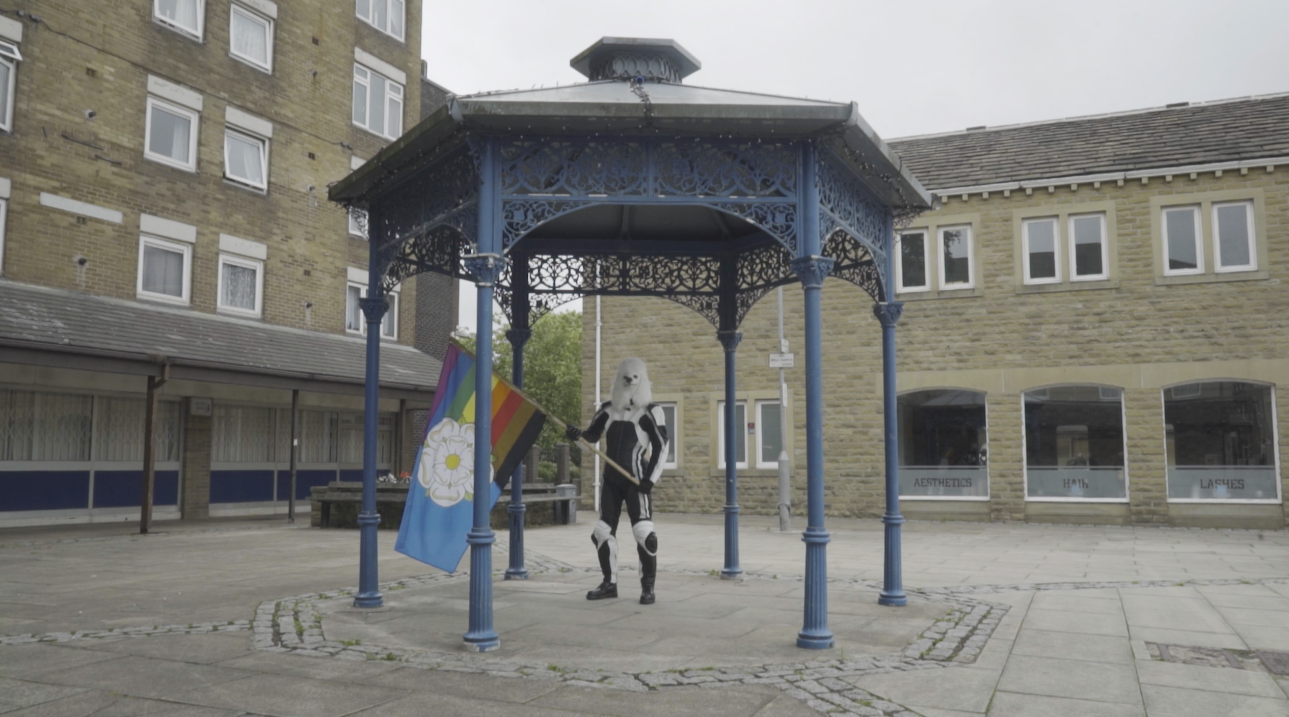 A humanoid dog waves a Yorkshire rainbow flag from inside a bandstand